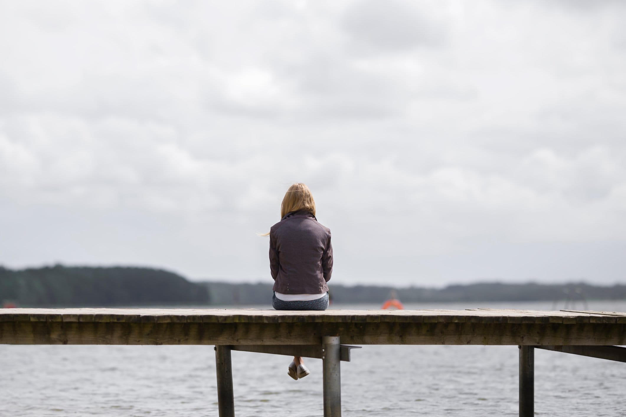One young woman sitting alone on edge of footbridge and staring at lake and cloudy sky in summer day. Thinking about life. Spending time alone in nature. Peaceful atmosphere. Back view.
